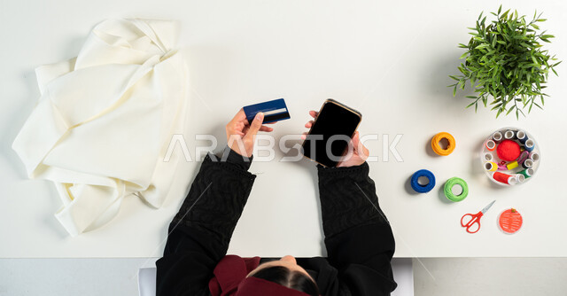 التسوق من خلال المواقع الإلكترونية، بورتريه ليدي إمرأة عربية خليجية سعودية تقوم باستخدام الجوال للقيام بالدفع عبر بطاقة المصرف البنكية وبجانبها مستلزمات الخياطة، البيع والشراء عبر الانترنت، الحصول على أدوات الخياطة أونلاين، عرض شاشة سوداء فارغة، خلفية بيضاء