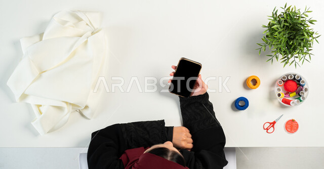 الاستفادة من المواقع الإلكترونية للتعلم والتدريب، بورتريه ليدي إمرأة عربية خليجية سعودية تقوم باستخدام الهاتف المحمول وبجانبها مستلزمات الخياطة، البيع والشراء عبر الانترنت، الحصول على أدوات الخياطة أونلاين، تفصيل الملابس والثياب، عرض شاشة سوداء فارغة، خلفية بيضاء