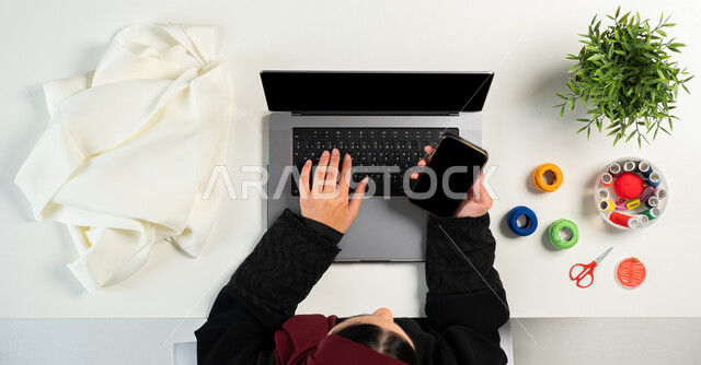 التعلم والتدريب من خلال المواقع الإلكترونية، بورتريه ليدي إمرأة عربية خليجية سعودية تقوم باستخدام اللابتوب والجوال بجانبها مستلزمات الخياطة، البيع والشراء عبر الانترنت، الحصول على أدوات الخياطة أونلاين، تفصيل الملابس والثياب، عرض شاشة سوداء فارغة، خلفية بيضاء