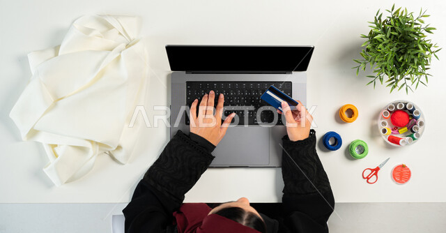 البيع والشراء عبر الانترنت، بورتريه ليدي إمرأة عربية خليجية سعودية تقوم باستخدام جهاز الحاسوب المحمول للدفع عبر بطاقة الصراف البنكية وبجانبها مستلزمات الخياطة، الحصول على الأقمشة وأدوات الخياطة أونلاين، تفصيل الملابس والثياب، عرض شاشة سوداء فارغة، خلفية بيضاء