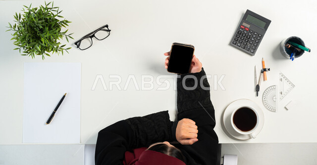 تعلم الحساب والرياضيات عبر المواقع الإلكترونية، بورتريه ليدي إمرأة عربية خليجية سعودية تقوم باستخدام الجوال وبجانبها أدوات هندسية وقرطاسية، البيع والشراء من خلال الانترنت، الحصول على المستلزمات الهندسية أونلاين، عرض شاشة سوداء فارغة، خلفية بيضاء