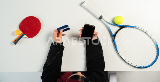 البيع والشراء الآمن، بورتريه ليدي إمرأة عربية خليجية سعودية تقوم باستخدام الهاتف المحمول للدفع عبر البطاقة البنكية وبجانبها أدوات رياضية، تعلم لعب رياضة التنس وكرة الطاولة عبر المواقع الالكترونية، تسوق المعدات الرياضية، عرض شاشة سوداء فارغة، خلفية بيضاء