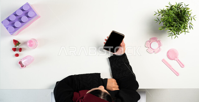 الحصول على المعلومات عبر الانترنت، البيع والشراء أونلاين، بورتريه مقرب ليدي إمرأة عربية خليجية سعودية تستخدم جهاز الهاتف المحمول وبجانبها ألعاب البيبي، التسوق من خلال المواقع الالكترونية، عرض شاشة سوداء فارغة، خلفية بيضاء