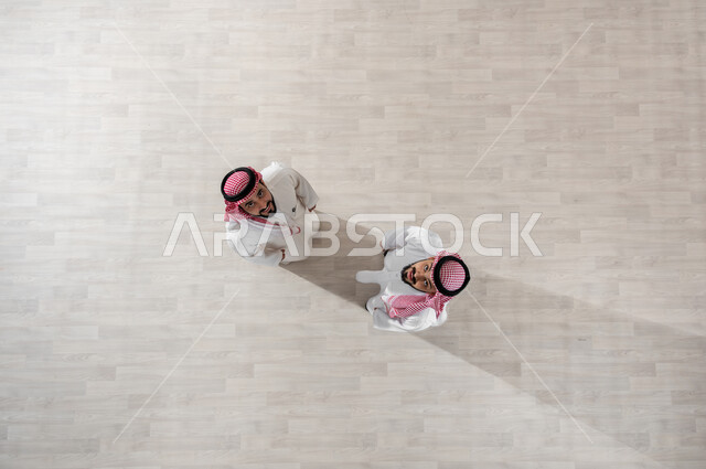 النظر الى الكاميرا بإيماءات الفرح والسرور, تعابير الصداقة الودية, قضاء أوقات ممتعة في العمل, التجول في ممرات الشركة, خروج الموظفين عند انتهاء أوقات الدوام الرسمية, بورتريه علوي عامودي لزملاء عمل عرب خليجيين سعوديين يرتديان الثوب التقليدي والشماغ, خلفية بيج