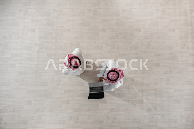 النظر الى الحاسب المحمول لرؤية شيء ما, استعمال التكنولوجيا في العمل, استخدام الأجهزة الحديثة في إنجاز مهام الأعمال, بورتريه علوي عامودي لزملاء عمل عرب خليجيين سعوديين يرتديان الثوب التقليدي والشماغ, خلفية بيج