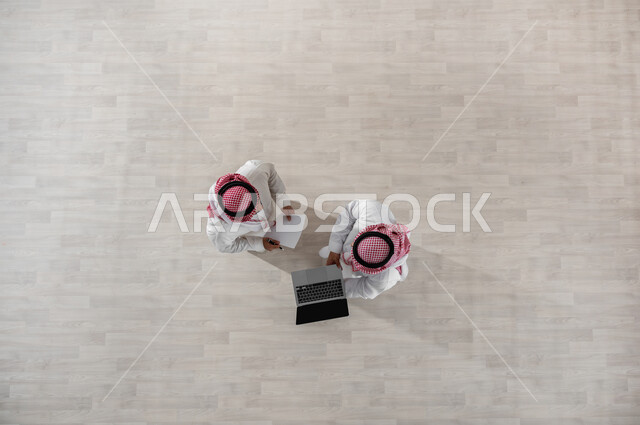 Using technology at work, using modern devices to complete business tasks, a vertical overhead portrait of Saudi Gulf Arab co-workers wearing the traditional thobe and shemagh, looking at the laptop to see something, beige background