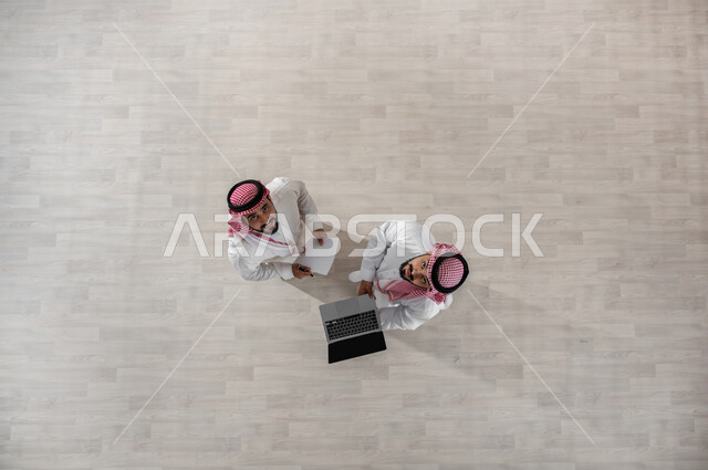 النظر الى الكاميرا بإيماءات الإنجاز والفرح, استخدام الأجهزة الحديثة في إنجاز مهام الأعمال, استعمال التكنولوجيا في العمل, بورتريه علوي عامودي لزملاء عمل عرب خليجيين سعوديين يرتديان الثوب التقليدي والشماغ, عرض شاشة سوداء فارغة على الحاسب المحمول, خلفية بيج