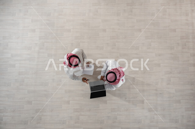 استخدام الأجهزة الحديثة في إنجاز مهام الأعمال, ااستعمال التكنولوجيا في العمل, بورتريه علوي عامودي لزملاء عمل عرب خليجيين سعوديين يرتديان الثوب التقليدي والشماغ,  الإشارة الى الحاسب المحمول بأصبع السبابة للدلالة على شيء ما, إنجاز العمل عبر التقنيات, خلفية بيج