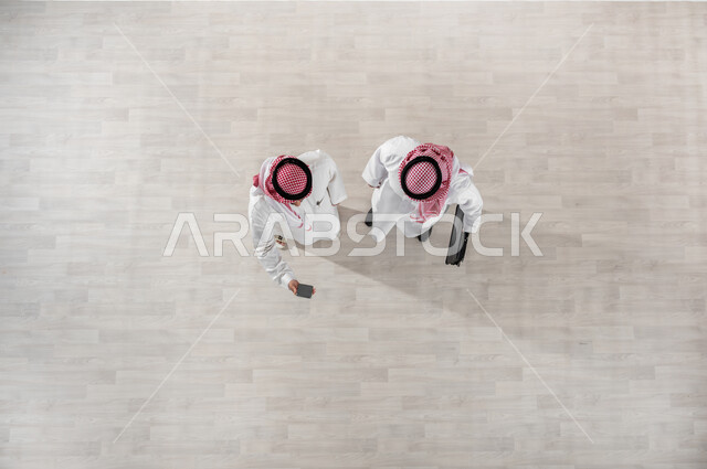 خروج الموظفين عند انتهاء أوقات الدوام الرسمية, استخدام التقنيات والتكنولوجيا الحديثة, التجول في ممرات الشركة, قضاء أوقات ممتعة في العمل, بورتريه علوي عامودي لزملاء عمل عرب خليجيين سعوديين يرتديان الثوب التقليدي والشماغ, النظر الى الهاتف المحمول, خلفية بيج