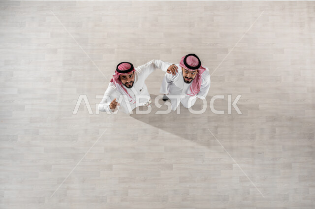 Walking around the company’s corridors, spending enjoyable times at work, employees leaving at the end of official working hours, a vertical overhead portrait of Saudi Gulf Arab co-workers wearing the traditional thobe and shemagh, looking at the camera with gestures of joy and pleasure, expressions of friendly friendship, beige background