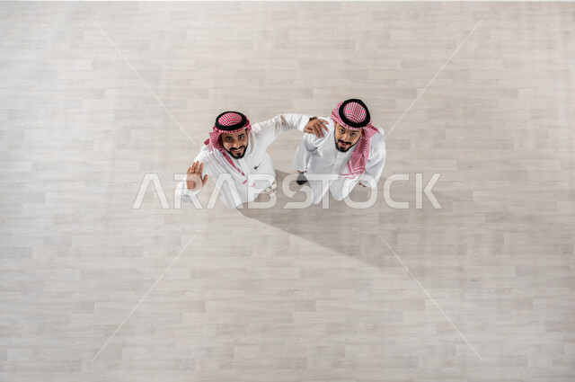 قضاء أوقات ممتعة في العمل, التجول في ممرات الشركة, خروج الموظفين عند انتهاء أوقات الدوام الرسمية, بورتريه علوي عامودي لزملاء عمل عرب خليجيين سعوديين يرتديان الثوب التقليدي والشماغ, النظر الى الكاميرا بإيماءات الفرح والسرور, تعابير الصداقة الودية, خلفية بيج