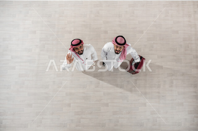 مفهوم السفر وزيارة مدن مختلفة, الاستعداد لبدء رحلة العمل بحماس, قضاء أوقات ممتعة في السياحة, بورتريه علوي عامودي لرجل عربي خليجي سعودي يرتدي الثوب التقليدي والشماغ يقوم بجر حقيبة سفر حمراء يقف مع صديقه, النظر الى الكاميرا بإيماءات السعادة والسرور, خلفية بيج