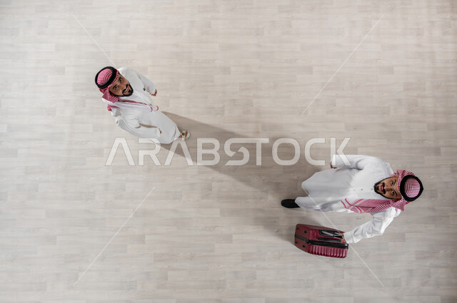 الاستعداد لبدء رحلة العمل بحماس, مفهوم السفر وزيارة مدن مختلفة, قضاء أوقات ممتعة في السياحة, بورتريه علوي عامودي لرجل عربي خليجي سعودي يرتدي الثوب التقليدي والشماغ يقوم بجر حقيبة سفر حمراء يقف مع صديقه, النظر الى الكاميرا بإيماءات السعادة والسرور, خلفية بيج