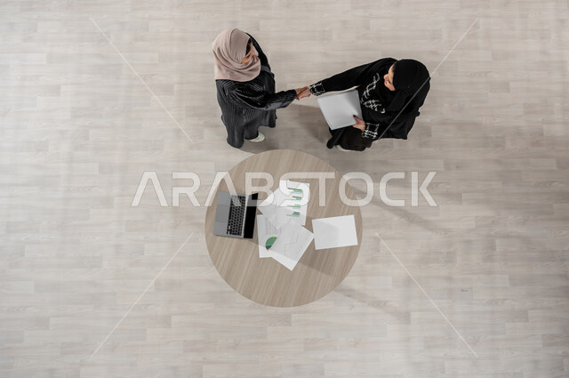 المصافحة والاتفاق في تطويرالاعمال, الاحترافية والتفاني في الانجاز, تبادل الخبرات والمعلومات الناجحة, التعاون والتفاهم بين الفريق, تعابير الصداقة العميقة, بورتريه علوي عامودي لسيدتين عربيتين خليجيتين سعوديتين في اجتماع, ارتداء اللباس السعودي التقليدي, خلفية بيج