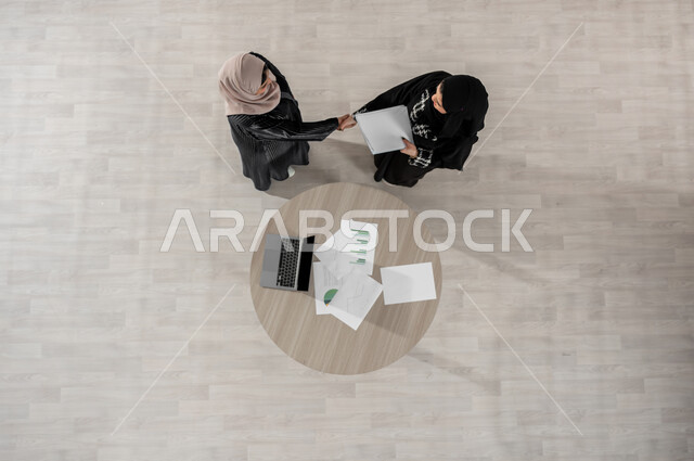 الاحترافية والتفاني في الانجاز, المصافحة والاتفاق في تطويرالاعمال, تبادل الخبرات والمعلومات الناجحة, التعاون والتفاهم بين الفريق, تعابير الصداقة العميقة, بورتريه علوي عامودي لسيدتين عربيتين خليجيتين سعوديتين في اجتماع, ارتداء اللباس السعودي التقليدي, خلفية بيج