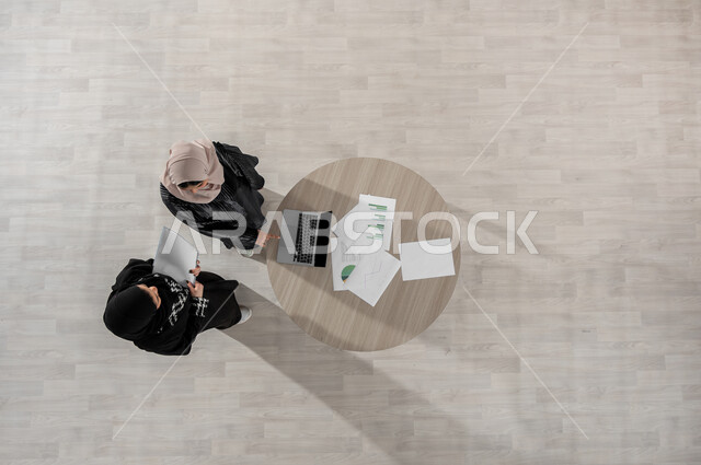 Cooperation and understanding between the team, exchange of experiences and successful agreements, vertical portrait of two Saudi Gulf Arab women in a business meeting, wearing traditional Saudi dress, comfortable work environment in the Saudi company, integrating technology into work life, beige background