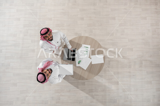 Looking at the camera with gestures of achievement, group interaction to achieve the company’s goals, a vertical overhead portrait of two Saudi Gulf Arab employees wearing the traditional shemagh and thobe, the concept of cooperative management, discussion of work matters using papers and data, integrating technology into work, beige background