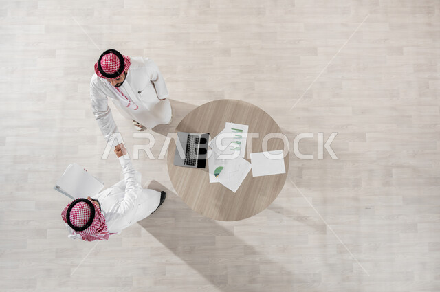 المصافحة والاتفاق في تطوير الأعمال، مفهوم الإدارة المتعاونة، دمج التكنولوجيا بالعمل، التفاعل الجماعي لتحقيق أهداف الشركة، بورتريه علوي عامودي لموظفين عربيين خليجيين سعوديين يرتديان الشماغ والثوب التقليدي، المناقشة بأمورالعمل باستخدام الأوراق والبيانات، خلفية بيج