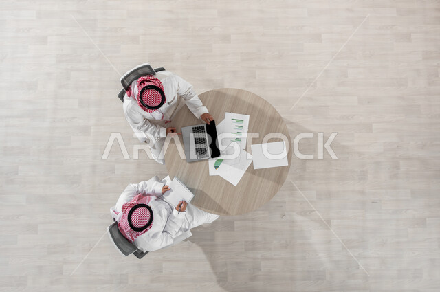 مفهوم الإدارة المتعاونة، دمج التكنولوجيا بالعمل، التفاعل الجماعي لتحقيق أهداف الشركة وحل المشكلات، بورتريه علوي عامودي لموظفين عربيين خليجيين سعوديين يرتديان الشماغ والثوب التقليدي، المناقشة في أمور العمل باستخدام الأوراق والبيانات، خلفية بيج