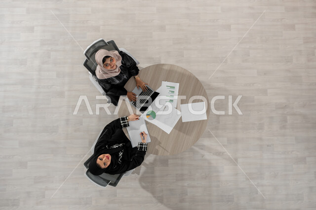 إنهاء الأعمال المكتبية في الوقت المحدد , بورتريه علوي عامودي لامرأتان عربيتان خليجيتان سعوديتان ترتديان الزي التقليدي , بيئة العمل في السعودية , النظر إلى الكاميرا بإيماءات السعادة و السرور  , عرض شاشة سوداء فارغة عبر الحاسوب , خلفية بيج
