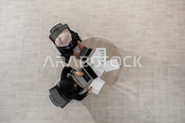 Illustration and explanation of the required tasks, a vertical overhead portrait of two Saudi Gulf Arab women wearing traditional clothing, the work environment in Saudi Arabia, using a laptop at work, displaying a blank black screen on the computer, beige background