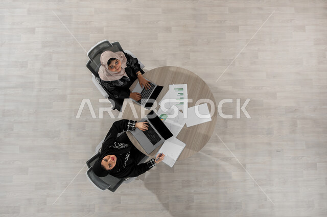 اكتساب الخبرات و المهارات الجديدة , بورتريه علوي عامودي لامرأتان عربيتان خليجيتان سعوديتان ترتديان الزي التقليدي , النظر إلى الكاميرا بإيماءات السعادة و السرور, استخدام جهاز الحاسب المحمول في العمل,  عرض شاشة سوداء فارغة عبر الحاسوب , خلفية بيج