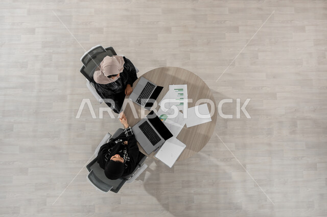 المصافحة و الاتفاق على المهام  , بورتريه علوي عامودي لامرأتان عربيتان خليجيتان سعوديتان ترتديان الزي التقليدي , دمج التكنولوجيا بالعمل , مناقشة و تقسيم المهام , عرض شاشة سوداء فارغة عبر الحاسوب , خلفية بيج