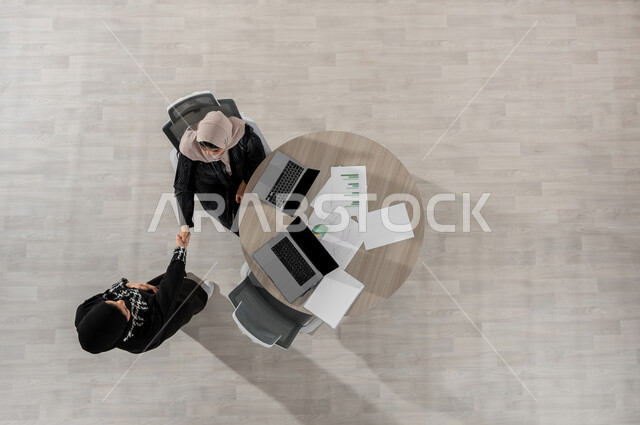 إجراء مقابلة عمل في مقر الشركة , بورتريه علوي عامودي لامرأتان عربيتان خليجيتان سعوديتان ترتديان الزي التقليدي , المصافحة و الاتفاق على المهام , دمج التكنولوجيا بالعمل , عرض شاشة سوداء فارغة عبر الحاسوب , خلفية بيج