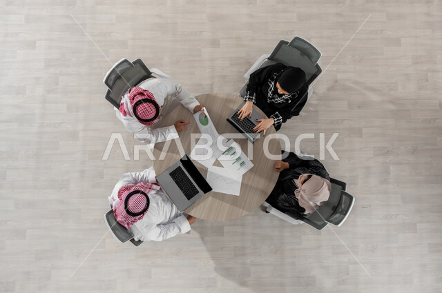 اجتماع عمل لمناقشة الاستراتيجيات و الأهداف , بورتريه علوي عامودي لفريق عمل عربي خليجي سعودي يرتدون الزي التقليدي , تنسيق و تقسيم المهام, استخدام جهاز الحاسب المحمول , عرض شاشة سوداء عبر الحاسوب , خلفية بيج