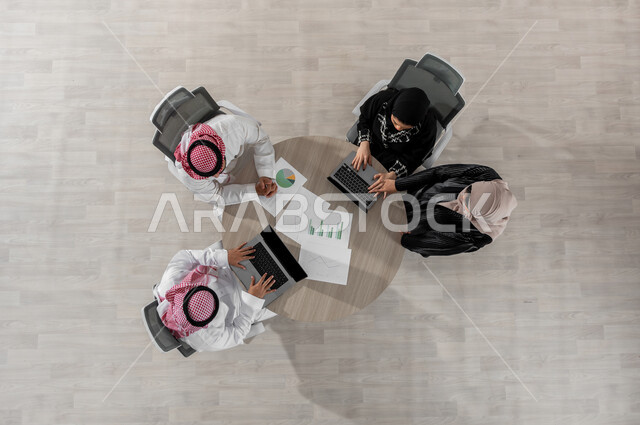 اجتماع عمل لشرح و تحليل البيانات , بورتريه علوي عامودي لفريق عمل عربي خليجي سعودي يرتدون الزي التقليدي , تطوير المشاريع و الاستثمارات , استخدام الحاسب المحمول في العمل , عرض شاشة سوداء عبر الحاسوب , خلفية بيج