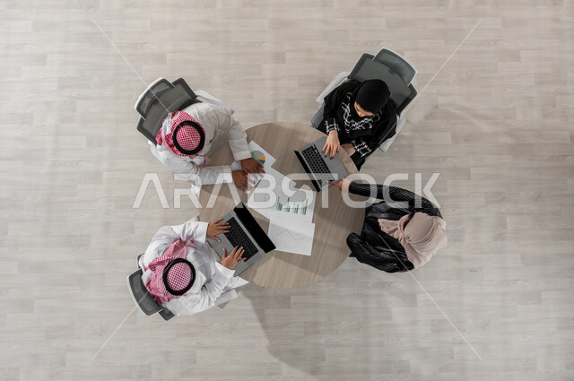 اجتماع عمل لدراسة أوراق البيانات الإحصائية , بورتريه علوي عامودي لفريق عمل عربي خليجي سعودي يرتدون الزي التقليدي , إدارة و تنسيق المهام , دمج التكنولوجيا بالعمل, عرض شاشة سوداء عبر الحاسوب , خلفية بيج