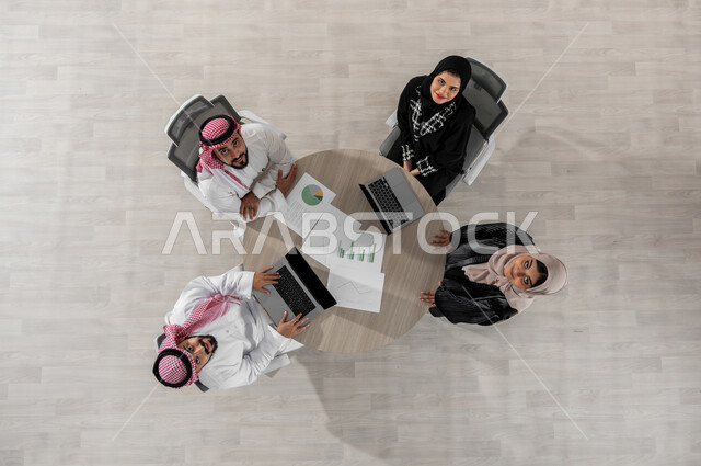 اجتماع عمل لتقسيم مهام المشاريع الجديدة , بورتريه علوي عامودي لفريق عمل عربي خليجي سعودي يرتدون الزي التقليدي , دمج التكنولوجيا بالعمل , النظر إلى الكاميرا بإيماءات الفرح و السرور , عرض شاشة سوداء عبر الحاسوب , خلفية بيج