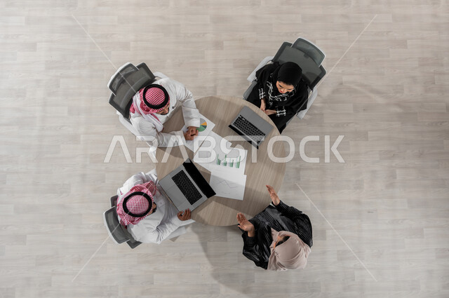 اجتماع عمل لتقسيم الأعمال المكتبية , بورتريه علوي عامودي لفريق عمل عربي خليجي سعودي يرتدون الزي التقليدي , دراسة و مراجعة المشاريع , استخدام جهاز الحاسب المحمول , عرض شاشة سوداء عبر الحاسوب , خلفية بيج