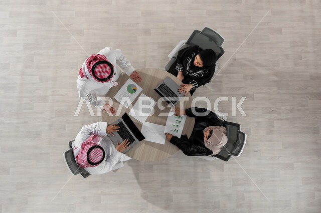 اجتماع عمل لمتابعة و مناقشة المشاريع , بورتريه علوي عامودي لفريق عمل عربي خليجي سعودي يرتدون الزي التقليدي , إدارة  وتنسيق المهام , دمج التكنولوجيا بالعمل , عرض شاشة سوداء عبر الحاسوب , خلفية بيج