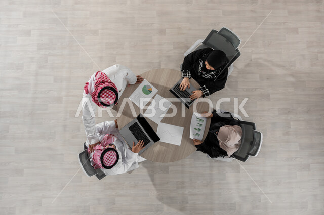 اجتماع عمل لمتابعة و مناقشة المهام , بورتريه علوي عامودي لفريق عمل عربي خليجي سعودي يرتدون الزي التقليدي , دمج التكنولوجيا بالعمل , إدارة و تنسيق المشاريع, عرض شاشة سوداء عبر الحاسوب , خلفية بيج