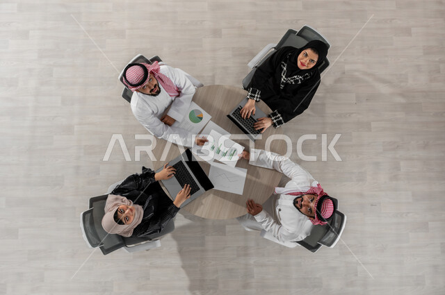 اجتماع عمل لتحليل بيانات الأعمال التجارية , بورتريه علوي عامودي لفريق عمل عربي خليجي سعودي يرتدون الزي التقليدي , تطوير المشاريع و الاستثمارات , النظر إلى الكاميرا بإيماءات السعادة والسرور , عرض شاشة سوداء عبر الحاسوب , خلفية بيج