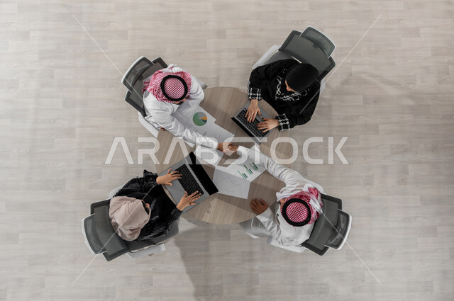 Shaking hands, concluding agreements and business deals, a vertical overhead portrait of a Saudi Gulf Arab work team wearing traditional clothing, a business meeting to develop projects and investments, a black screen displayed on the computer, a beige background