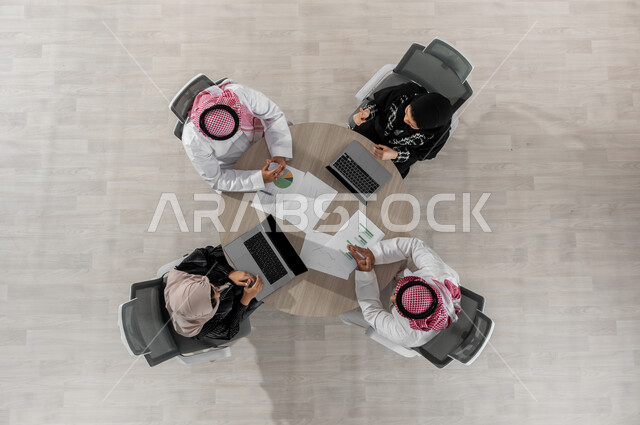 A business meeting to discuss tasks at the company’s headquarters, a vertical overhead portrait of a Saudi Gulf Arab work team wearing traditional clothing, studying and analyzing graphs of company profits, displaying a black screen on the computer, beige background
