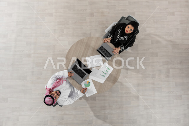 مراجعة و تدقيق المهام , النظر إلى الكاميرا بإيماءات السعادة و السرور , بورتريه علوي عامودي لرجل و سيدة أعمال عربيان خليجيان سعوديان يرتديان الزي التقليدي , مفهوم الإدارة و التنسيق, عرض شاشة سوداء عبر الحاسوب , خلفية بيج