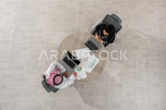 Exchanging experiences and skills, vertical portrait of a Saudi Gulf Arab man and businesswoman wearing traditional clothing, analyzing and understanding statistical data sheets, displaying a black screen via the computer, beige background