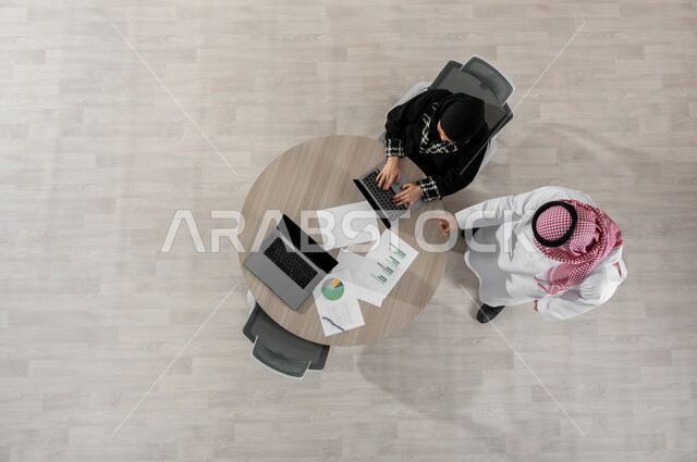 الإشارة بإصبع السبابة إلى جهاز الحاسب المحمول , بورتريه علوي عامودي لرجل و سيدة أعمال عربيان خليجيان سعوديان يرتديان الزي التقليدي , استخدام الأجهزة التقنية في إنجاز المهام ,عرض شاشة سوداء عبر الحاسوب , خلفية بيج