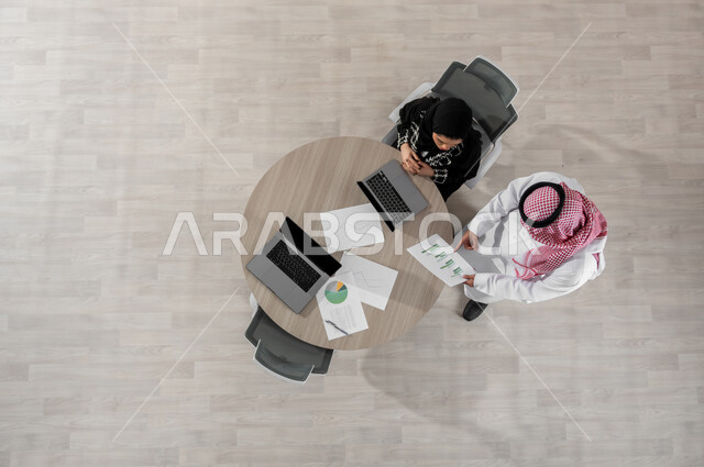 Managing and coordinating tasks, vertical overhead portrait of a Saudi Arabian Gulf businessman and woman wearing traditional clothing, reviewing projects and office work, pointing with the index finger at data sheets, displaying a black screen on the computer, beige background