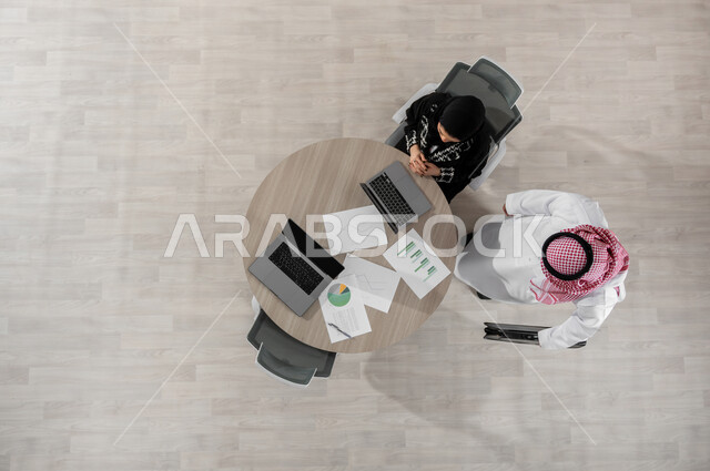 مراجعة المشاريع و الأعمال المكتبية , بورتريه علوي عامودي لرجل و سيدة أعمال عربيان خليجيان سعوديان يرتديان الزي التقليدي , إدارة و تنسيق المهام, عرض شاشة سوداء عبر الحاسوب , خلفية بيج