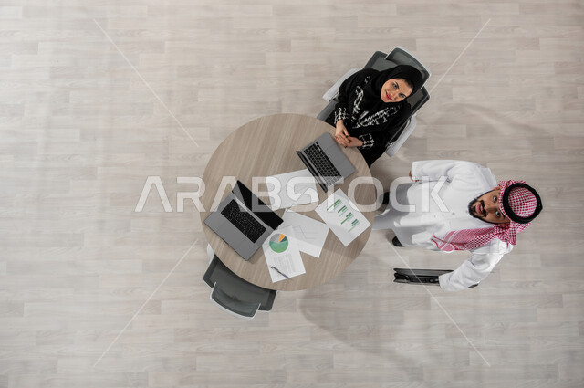 القيام بالأعمال المكتبية , بورتريه علوي عامودي لرجل و سيدة أعمال عربيان خليجيان سعوديان يرتديان الزي التقليدي , النظر إلى الكاميرا بإيماءات السعادة و السرور, عرض شاشة سوداء عبر الحاسوب , خلفية بيج