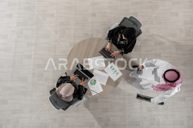 مناقشة و تنظيم الأعمال ,اجتماع عمل لشرح و تحليل البيانات الإحصائية , بورتريه علوي عامودي لفريق عمل عربي خليجي سعودي يرتدون الزي التقليدي, استخدام التقنيات الحديثة في إنجاز المهام , عرض شاشة سوداء عبر الحاسوب , خلفية بيج