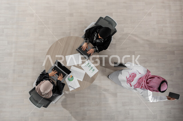 Using modern technologies to accomplish tasks, exchanging ideas and information, a vertical overhead portrait of a Saudi Gulf Arab work team wearing traditional clothing, a business meeting to discuss and divide work, displaying a black screen via computer and mobile phone, beige background