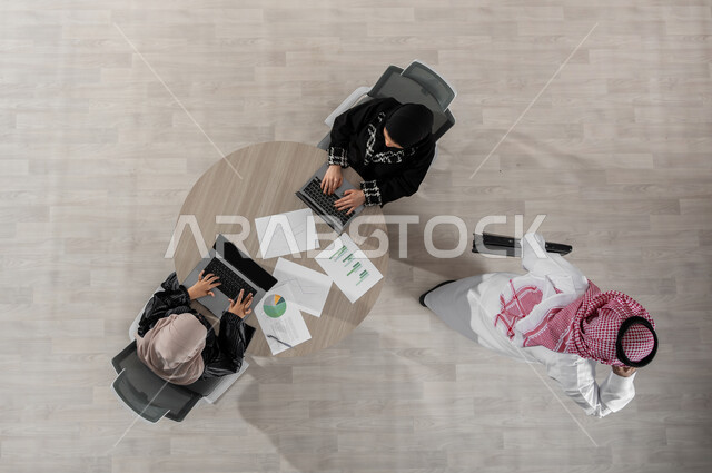 اجتماع عمل لتبادل الأفكار و المعلومات , استخدام التقنيات الحديثة في إنجاز المهام , بورتريه علوي عامودي لفريق عمل عربي خليجي سعودي يرتدون الزي التقليدي , مناقشة وتقسيم الأعمال, عرض شاشة سوداء عبر الحاسوب , خلفية بيج