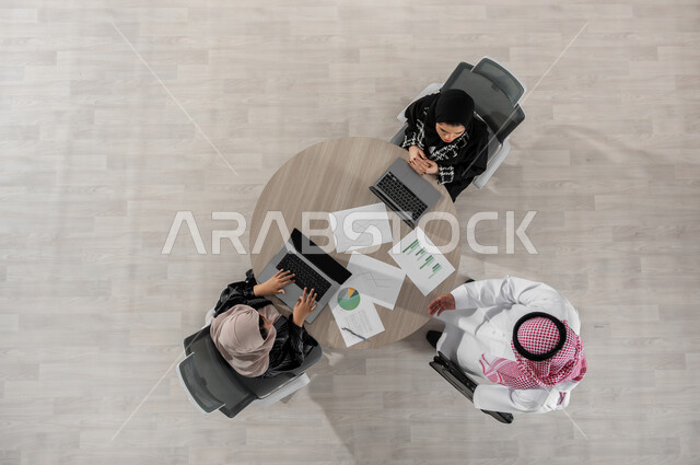 اجتماع عمل لشرح و تحليل البيانات الإحصائية , مناقشة و تنظيم الأعمال,  بورتريه علوي عامودي لفريق عمل عربي خليجي سعودي يرتدون الزي التقليدي, استخدام التقنيات الحديثة في إنجاز المهام  , عرض شاشة سوداء عبر الحاسوب , خلفية بيج