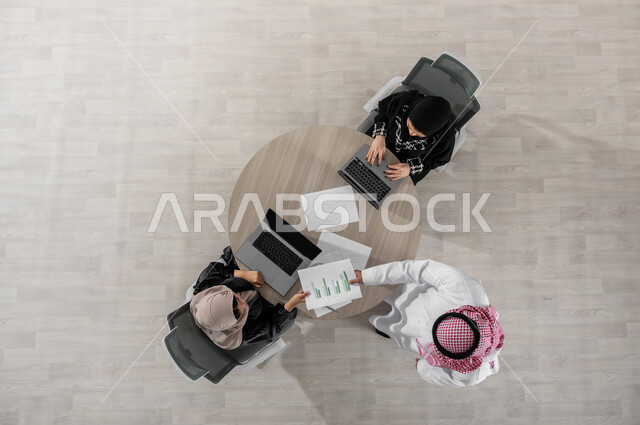 اجتماع عمل لمناقشة وتقسيم الأعمال , تبادل الأفكار و المعلومات , بورتريه علوي عامودي لفريق عمل عربي خليجي سعودي يرتدون الزي التقليدي , استخدام التقنيات الحديثة في إنجاز المهام , عرض شاشة سوداء عبر الحاسوب , خلفية بيج