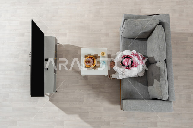 إيماءات التركيز و الاندماج , بورتريه علوي عامودي لرجل عربي خليجي سعودي يرتدي الزي التقليدي مع الشماغ  يجلس على الأريكة في غرفة المعيشة , قضاء أوقات الفراغ بمشاهدة البرامج التلفزيونية , عرض شاشة سوداء فارغة عبر التلفاز, خلفية بيج